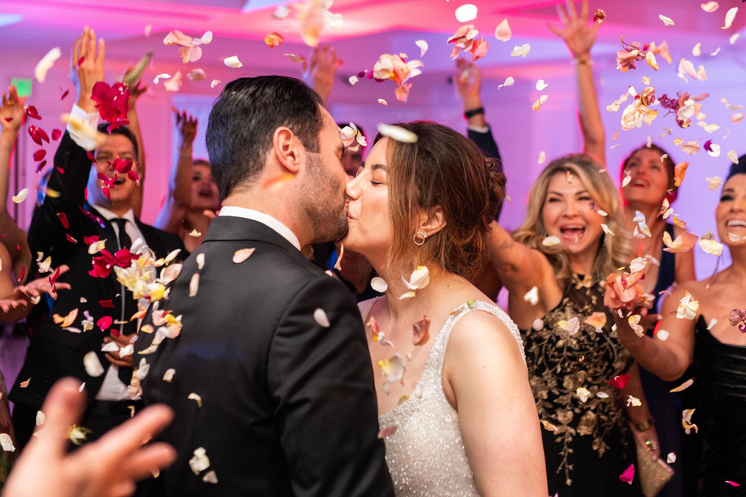 Newlyweds share a kiss on the dance floor surrounded by cheering guests throwing colorful confetti at their wedding reception.