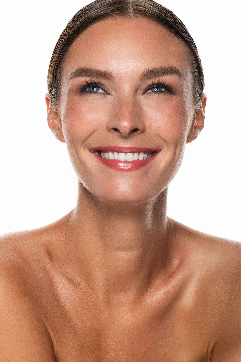 Smiling woman with glowing skin, bare shoulders, and blue eyes against a white background.
