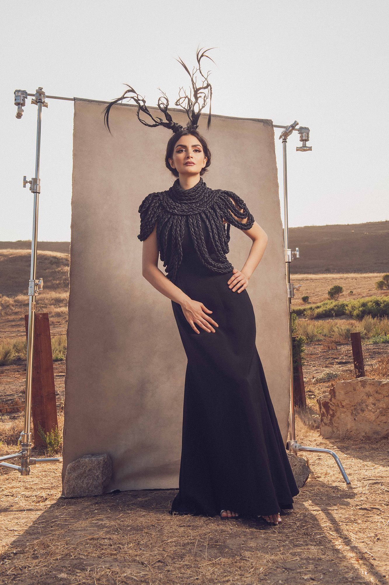 Woman in elegant black gown with ornate antler crown posing before outdoor photography backdrop.