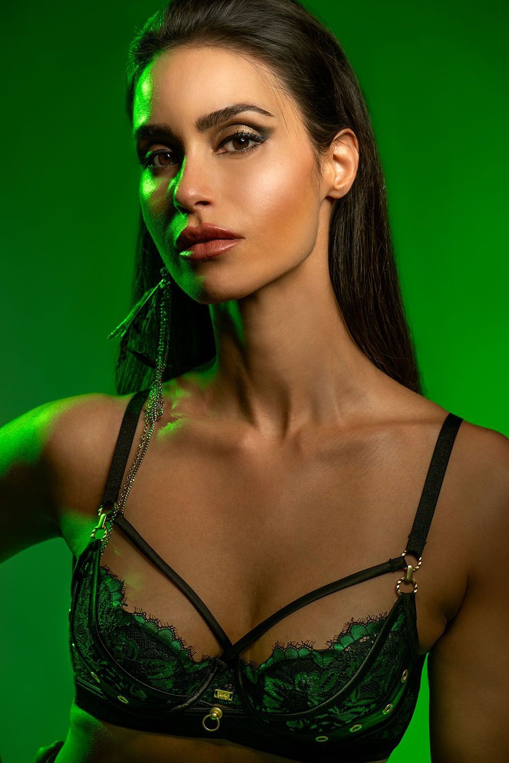 Glamorous brunette in black green lace bralette with feather earrings in green-lit studio.