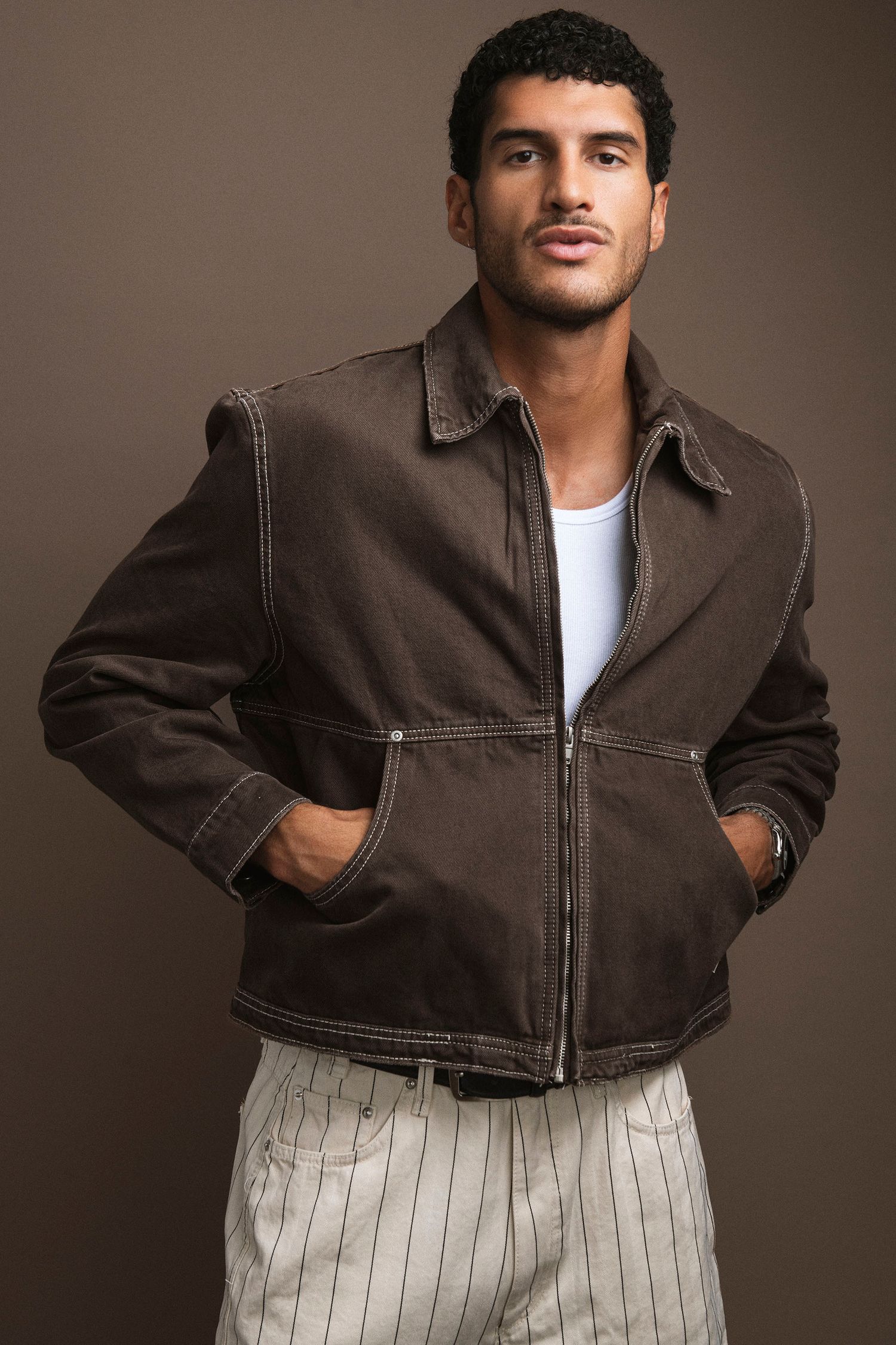 Man in brown zip-up jacket and striped trousers posing confidently in a studio.