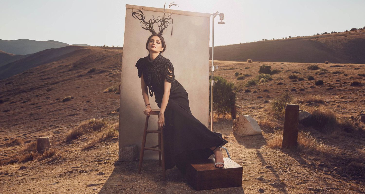 Woman in black dress sits on stool in desert landscape with surreal deer antler backdrop painting.