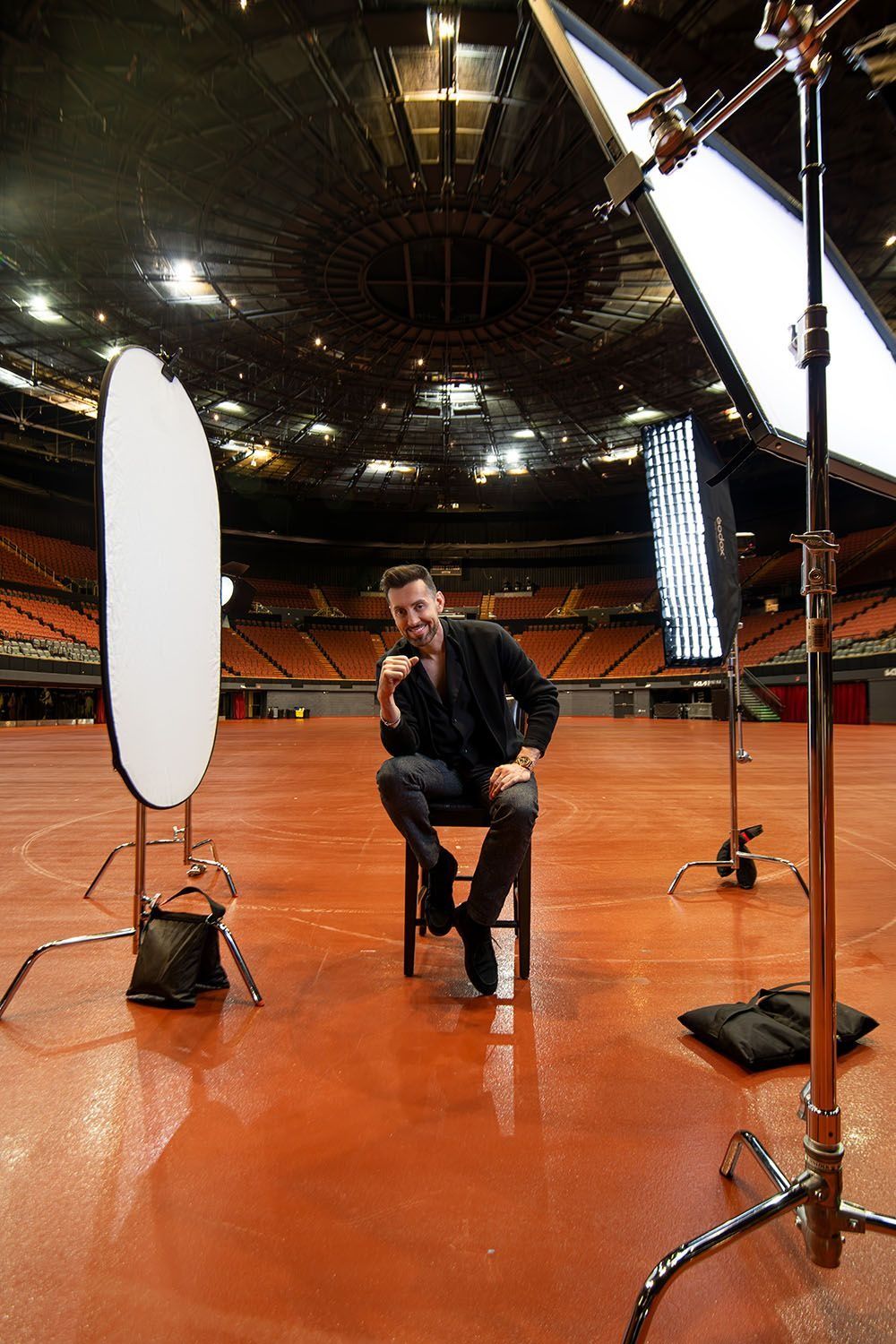 Man sitting on a stool surrounded by studio photography lights inside a large empty arena.