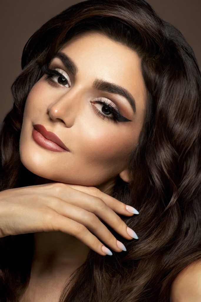 Beautiful woman with glamorous makeup, dark wavy hair, and white French tip nails posing in studio.