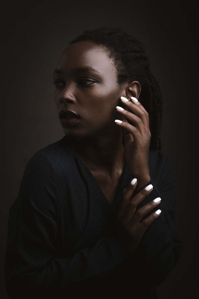Dramatic low-key portrait of a Black woman with braids touching her face against dark background.
