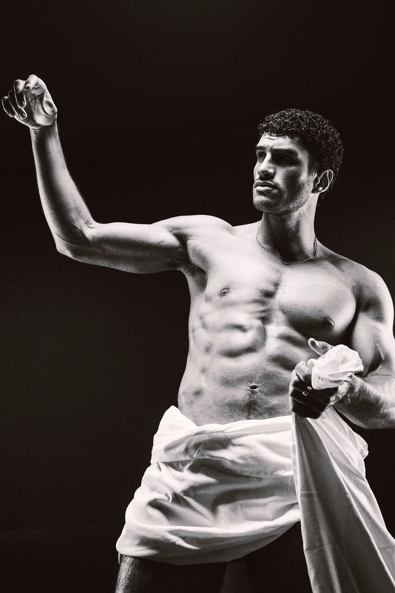 Black and white photo of a muscular shirtless man raising his fist, holding white fabric in a studio.