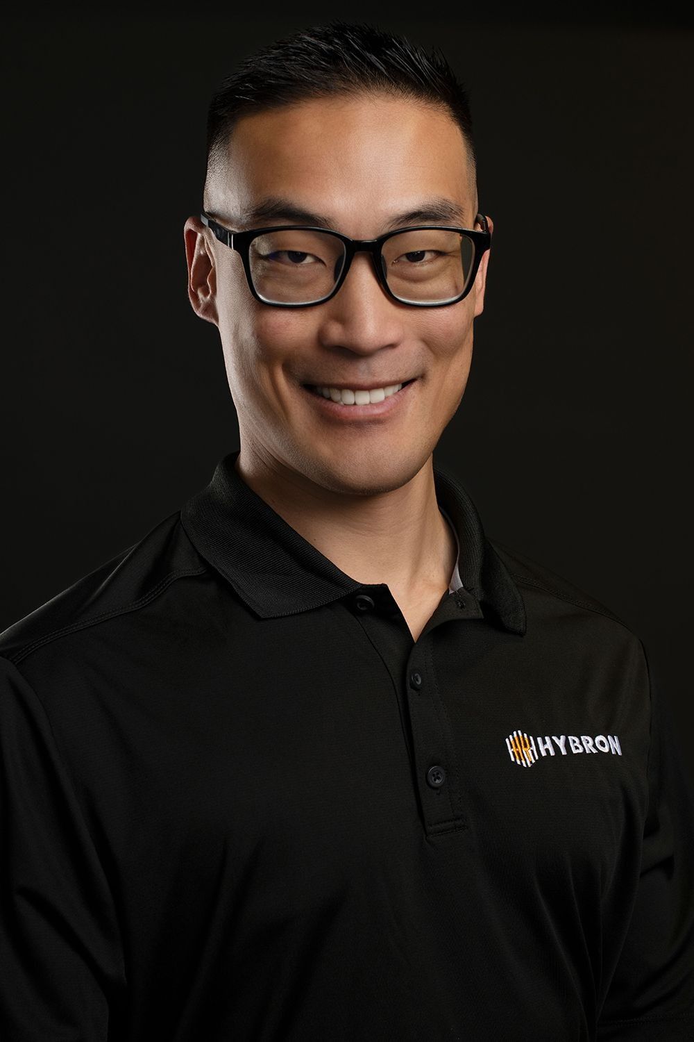 Professional man in black Hybron polo shirt smiles confidently against dark studio background.