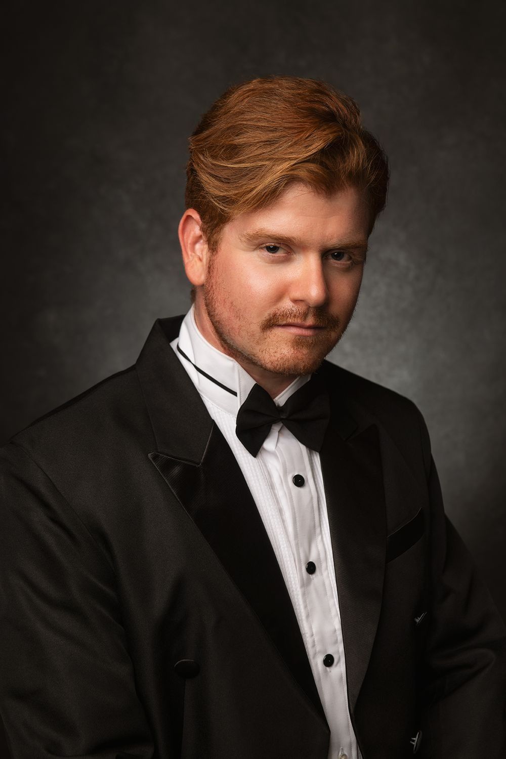 Elegant man with red hair wearing a black tuxedo and bow tie in a formal portrait.