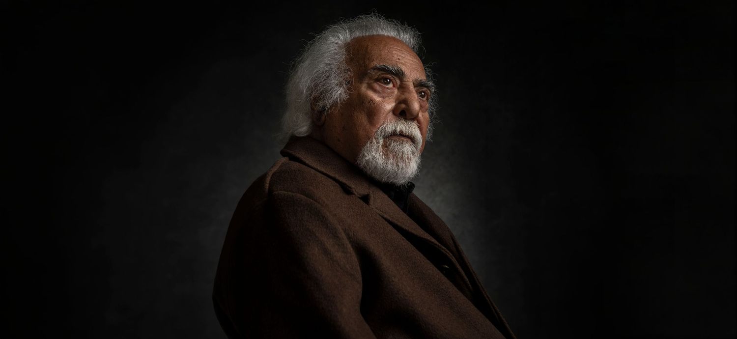 Elderly man with white hair and beard in brown jacket against dark background.