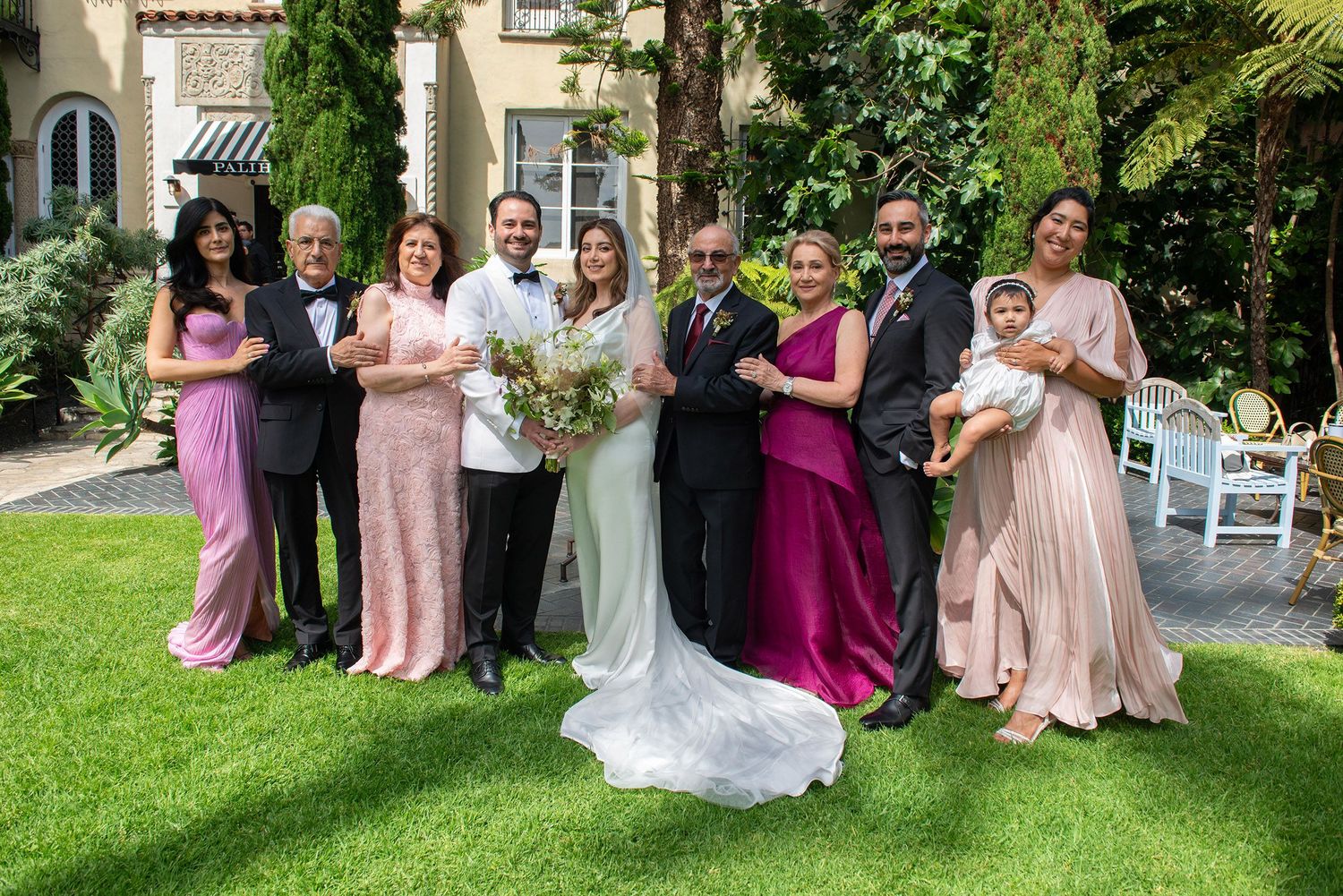 Wedding party poses outdoors on green lawn with bride in white gown and guests in pink and magenta formal attire.