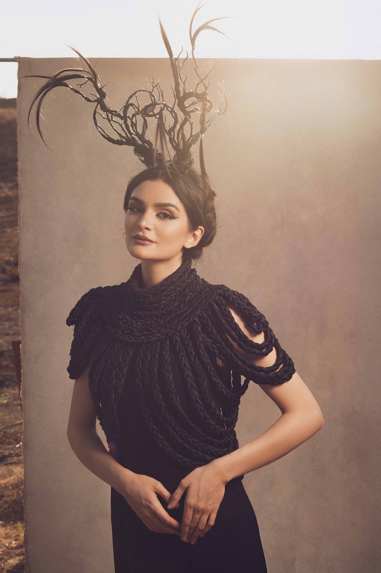 Model wearing black textured top with dramatic antler headpiece posing outdoors against neutral backdrop.