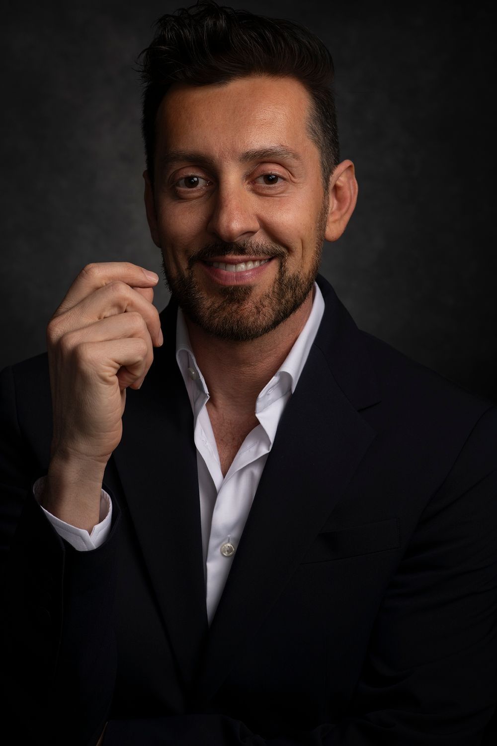 Smiling businessman in a black suit with open white shirt posing against a dark backdrop.