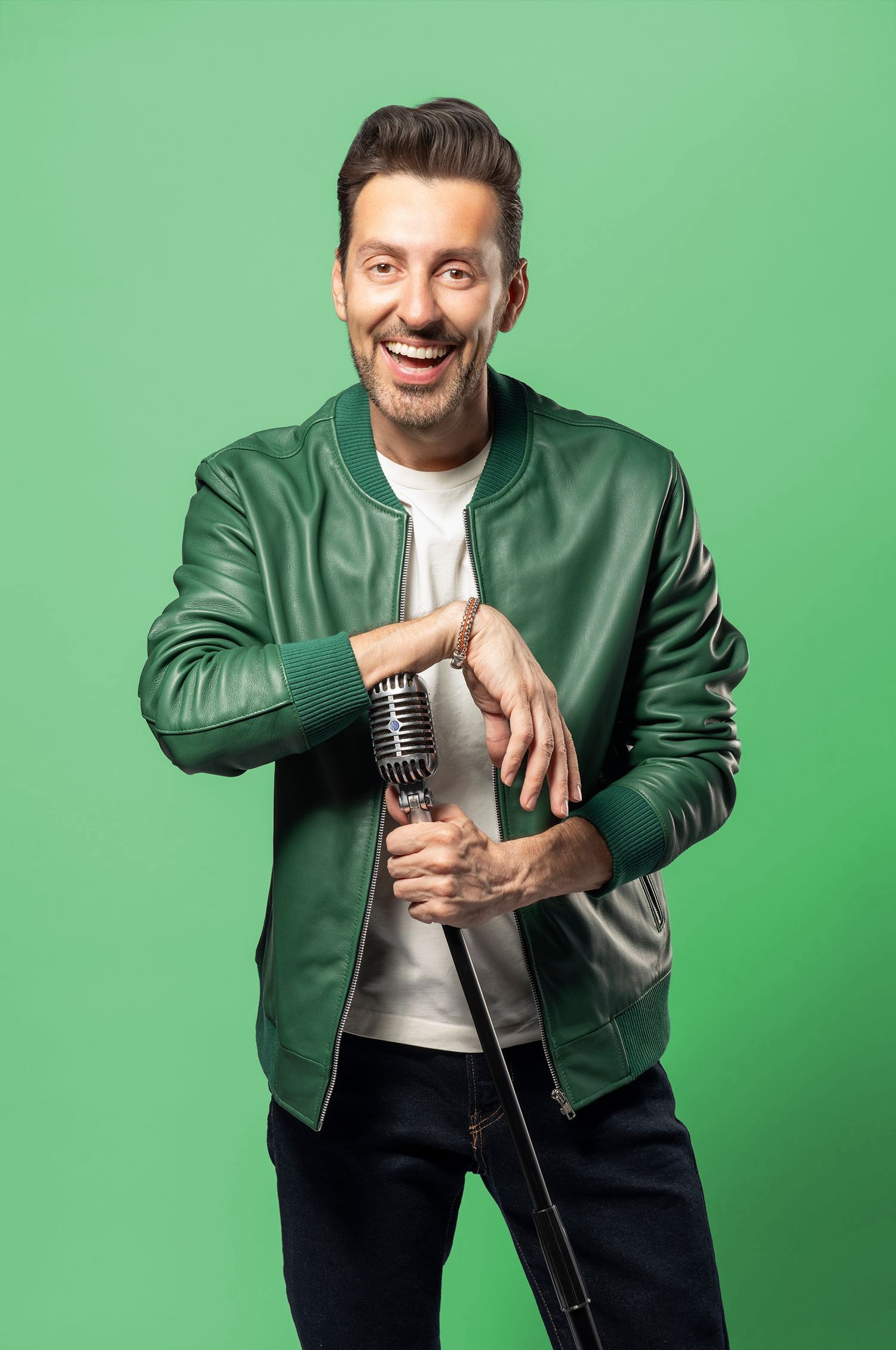 Smiling man in green leather jacket holding vintage microphone against bright green background.