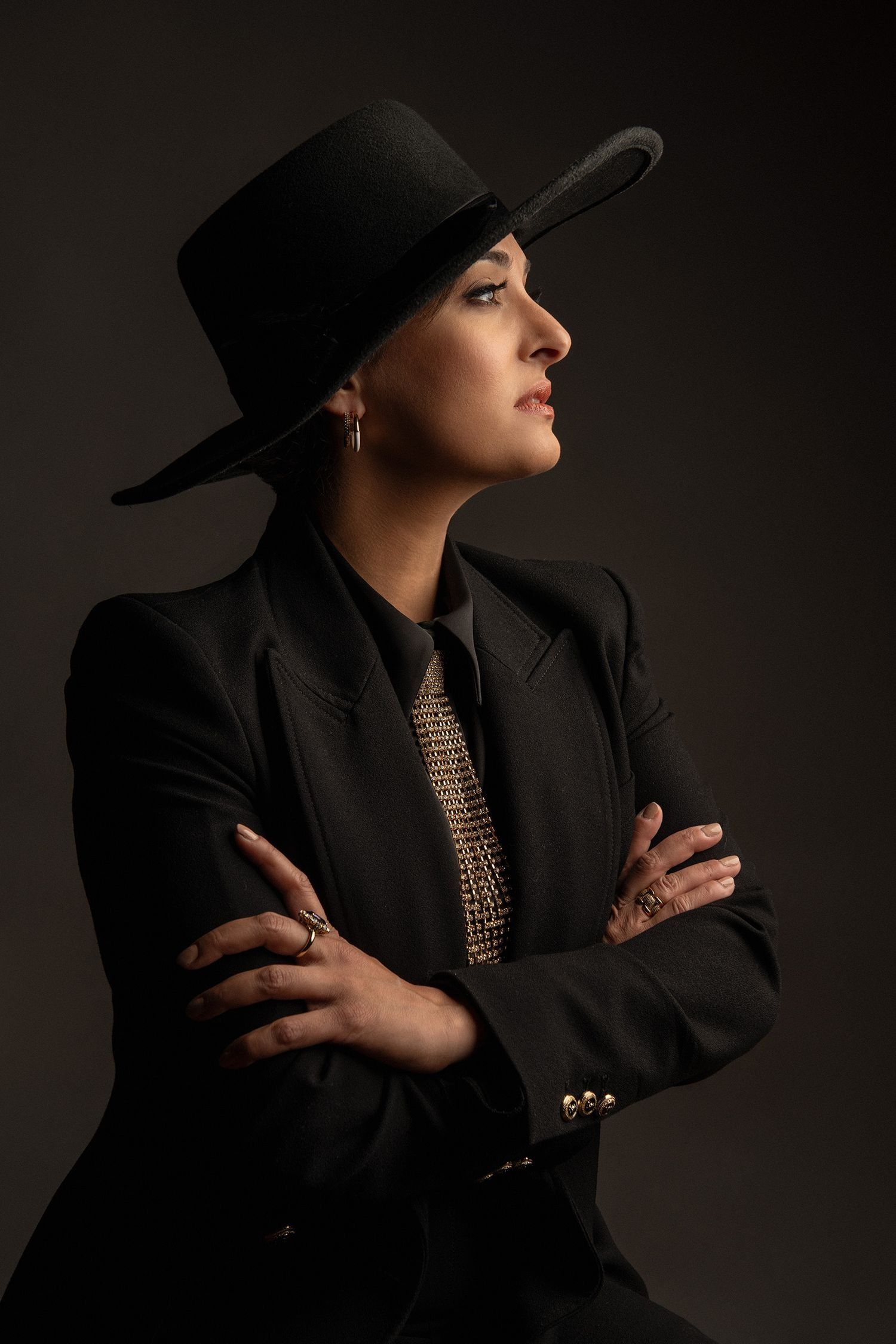 Elegant woman in black blazer and wide-brim hat with arms crossed, studio portrait.
