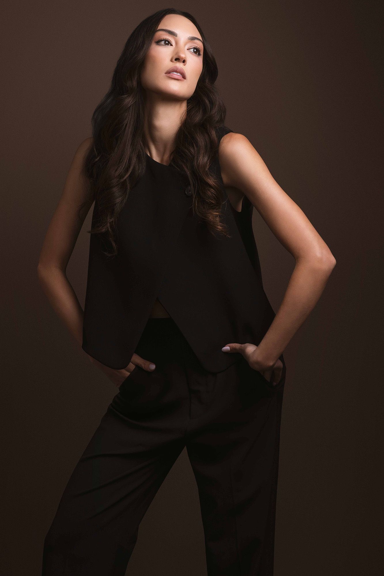 Female model in a sleeveless black wide-leg pantsuit posing with hands on hips on brown backdrop.