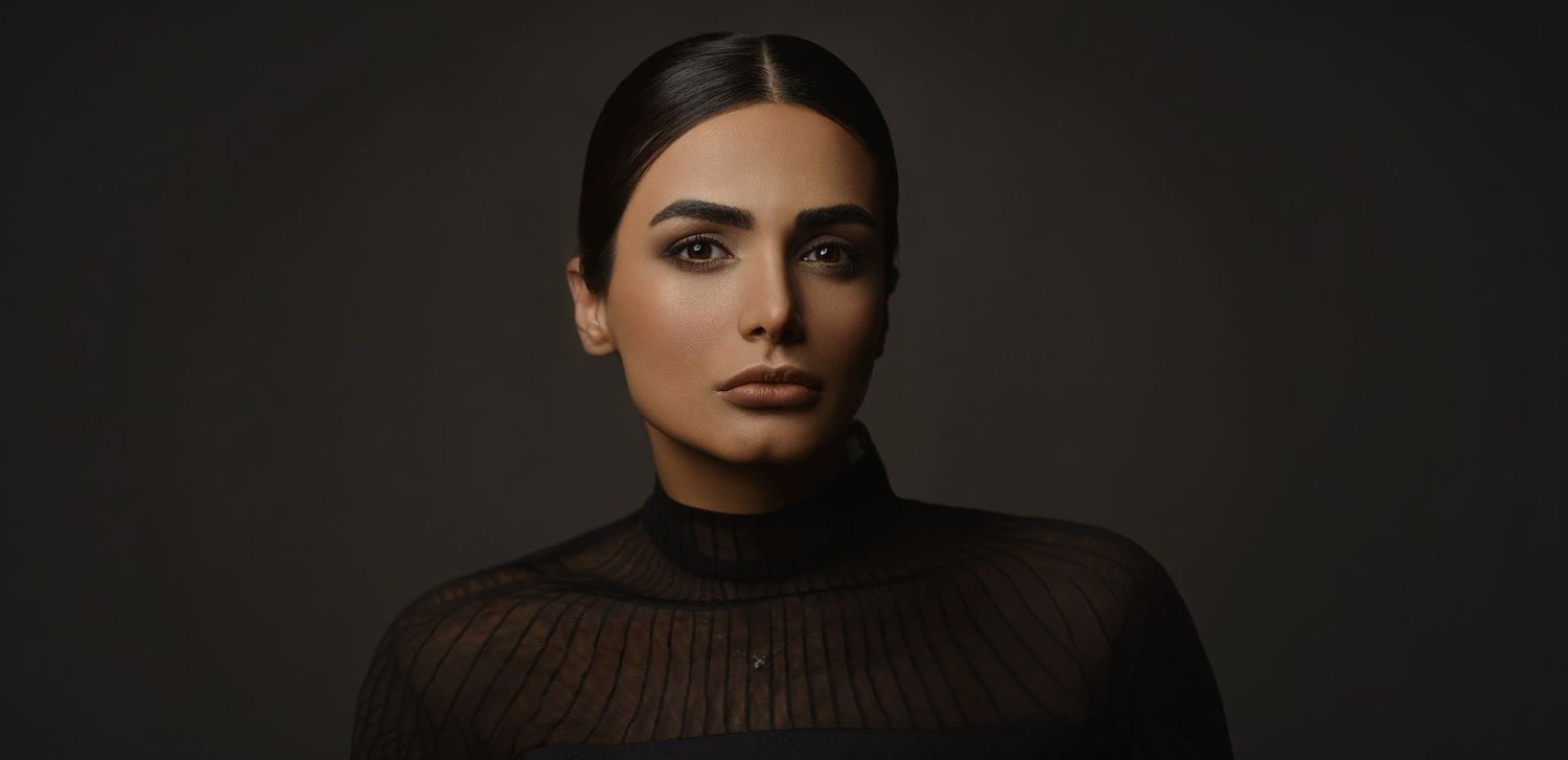 Woman with sleek dark hair in sheer black turtleneck giving intense gaze against dark moody background.