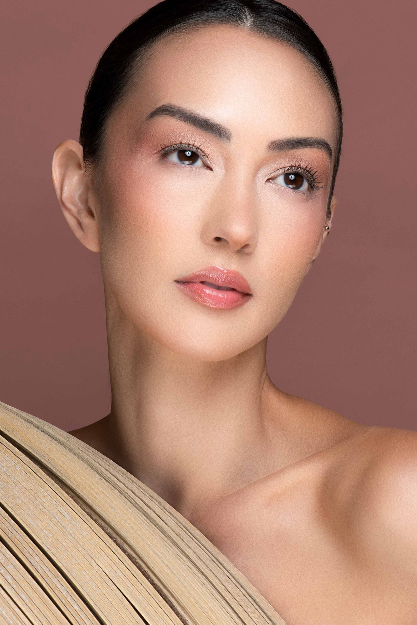 Asian woman with natural makeup posing with a woven fan accessory against a warm brown background.
