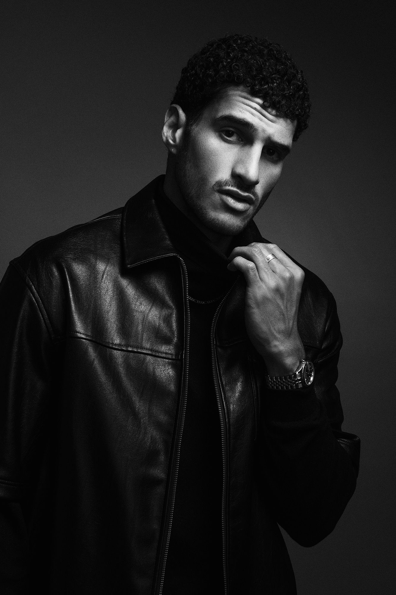 Black and white portrait of young man in leather jacket holding collar with watch on wrist.