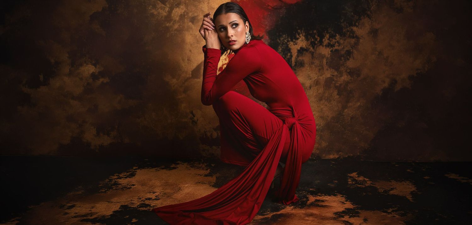 Woman in stunning red draped dress crouches gracefully against rich textured golden-brown background.