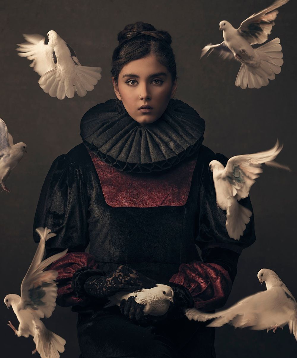 Young girl in Renaissance costume with white doves flying around her in a dramatic studio portrait.