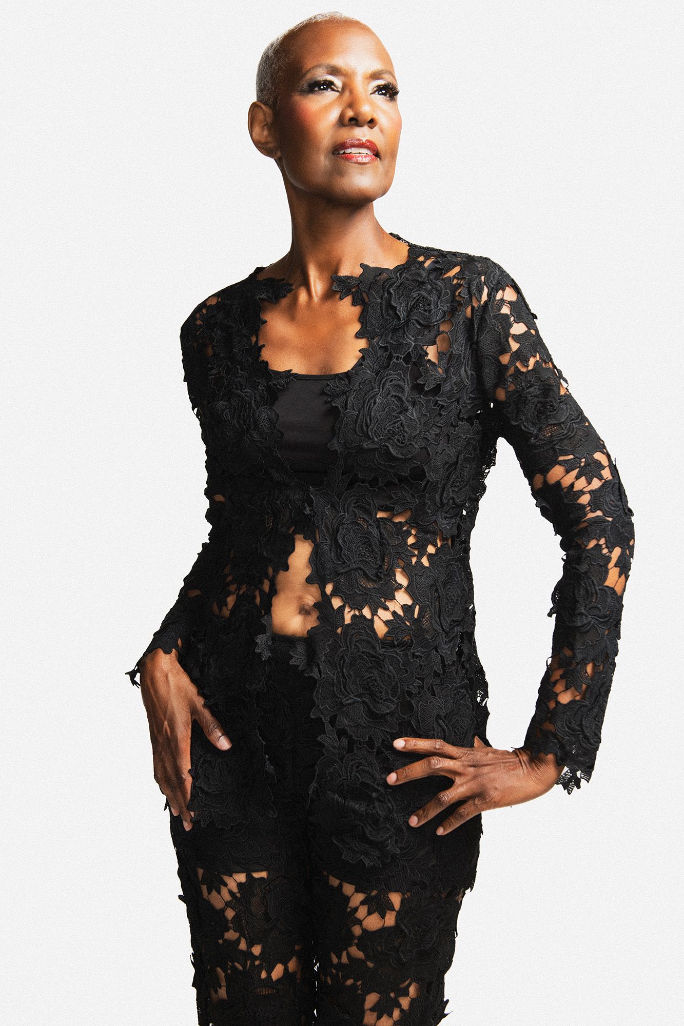 Elegant bald Black woman in black lace outfit posing confidently against white background.