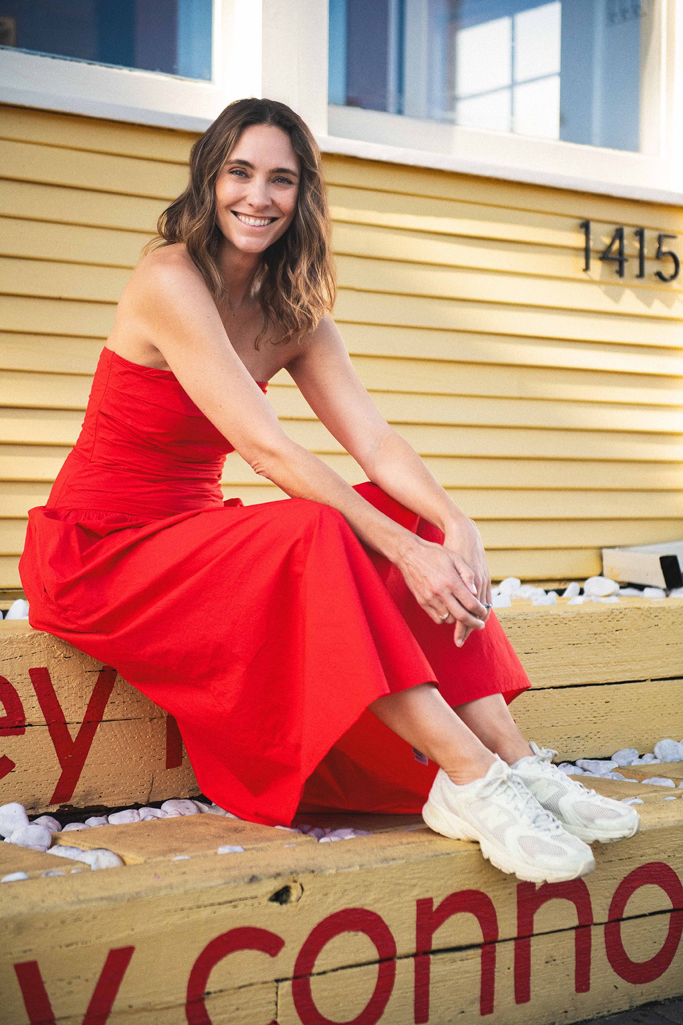 Woman in red strapless dress and white sneakers sitting outdoors smiling, posed against a yellow building.