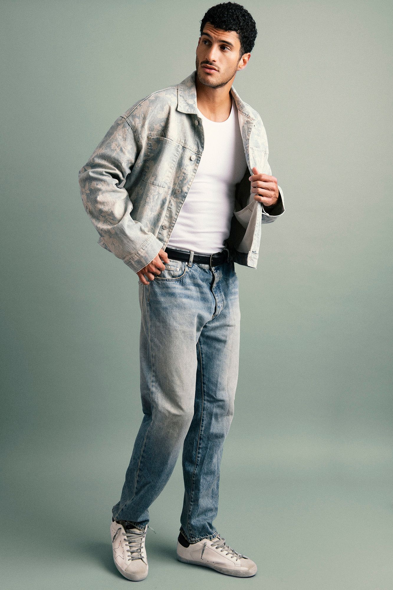 Man in gray denim jacket, white tee, light blue jeans, and white sneakers posing against a gray background.