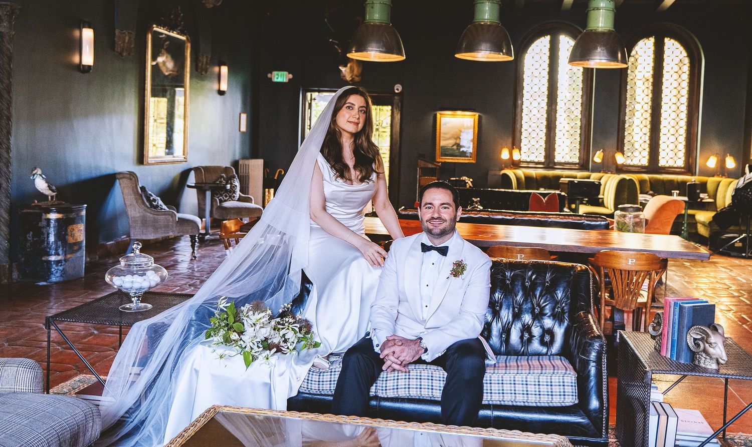 Bride in white gown and groom in tuxedo pose elegantly on a vintage sofa in a luxurious wedding venue.