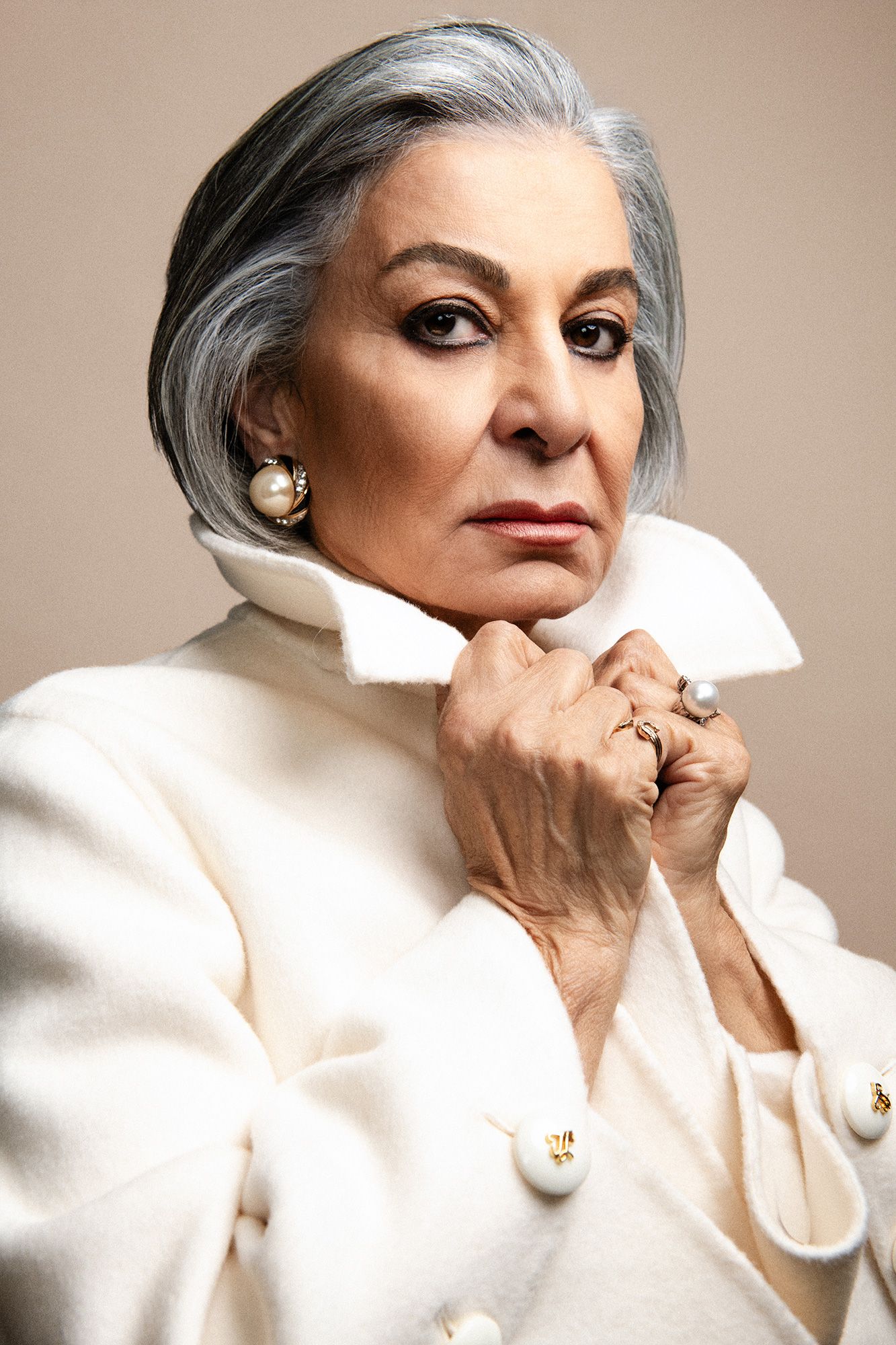 Elegant older woman with gray hair in white coat, pearl earrings, and rings, posing confidently against neutral background.