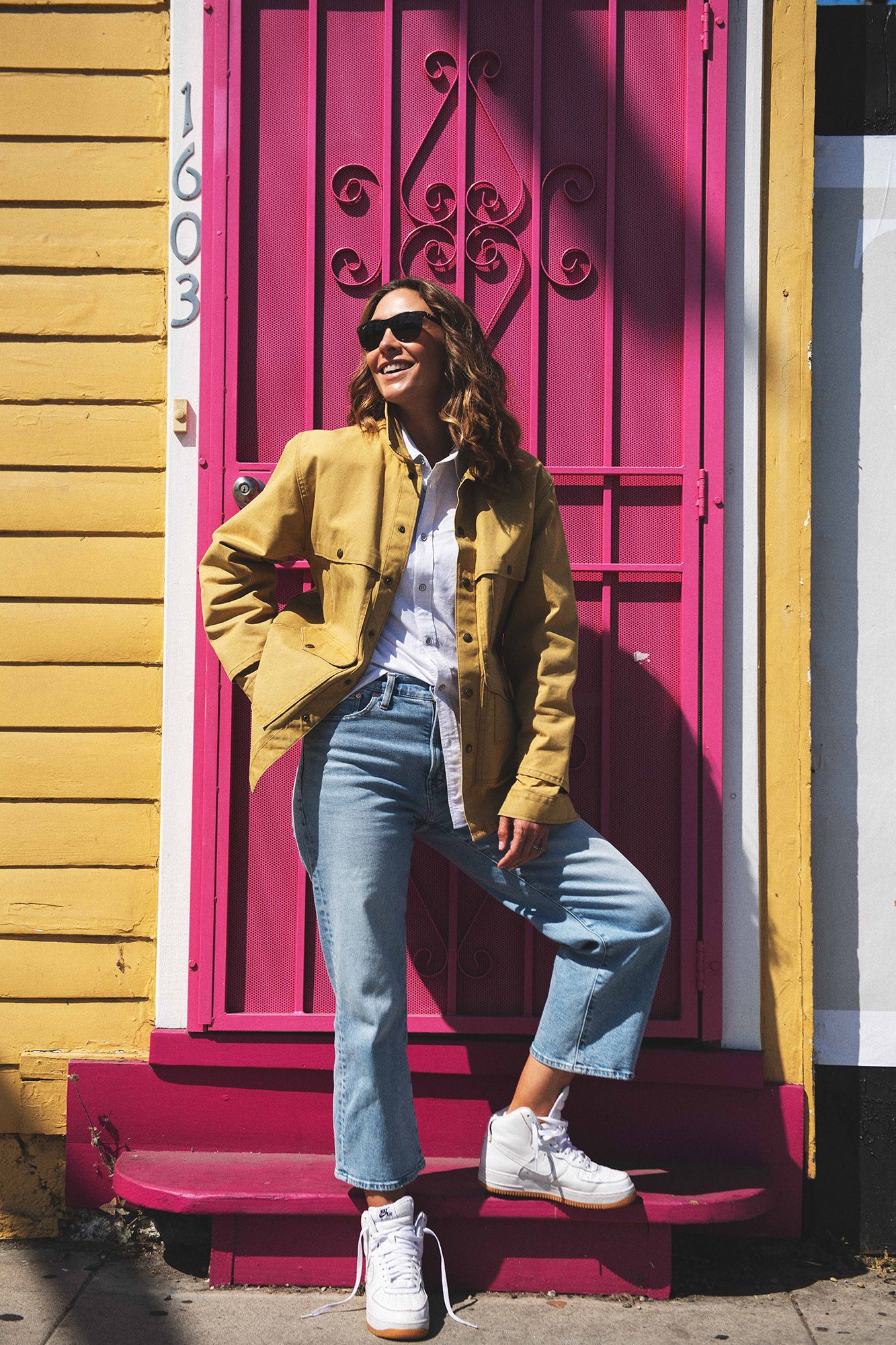 Woman in yellow jacket, jeans, and white sneakers smiling against a vibrant pink door.