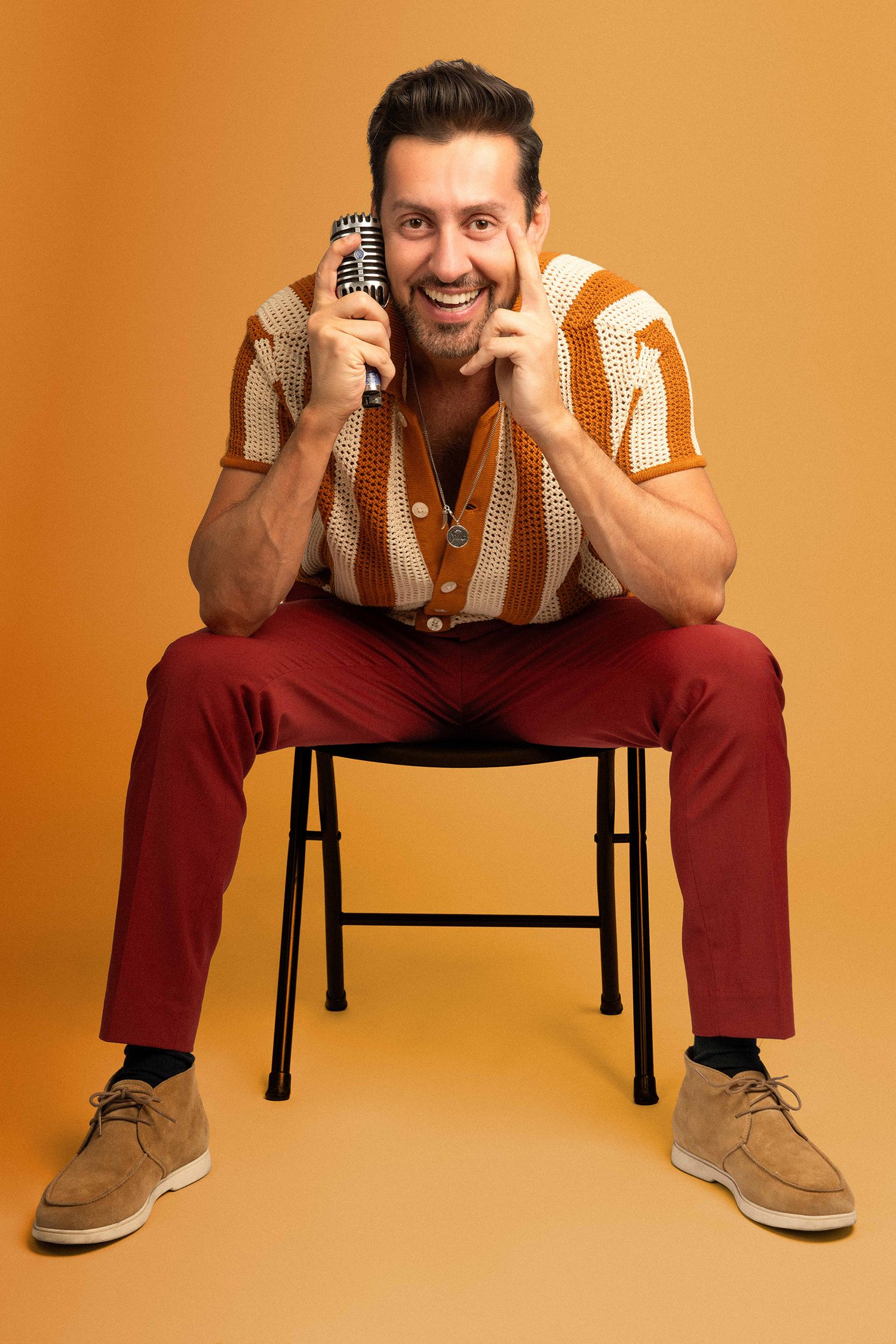 Smiling man sitting on chair holding vintage microphone, wearing striped shirt and red pants on orange background.
