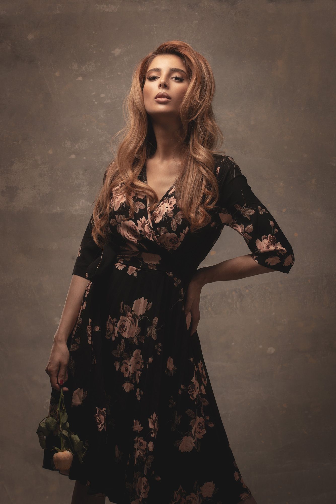 Blonde woman in black floral midi dress holding roses posing against textured vintage background.