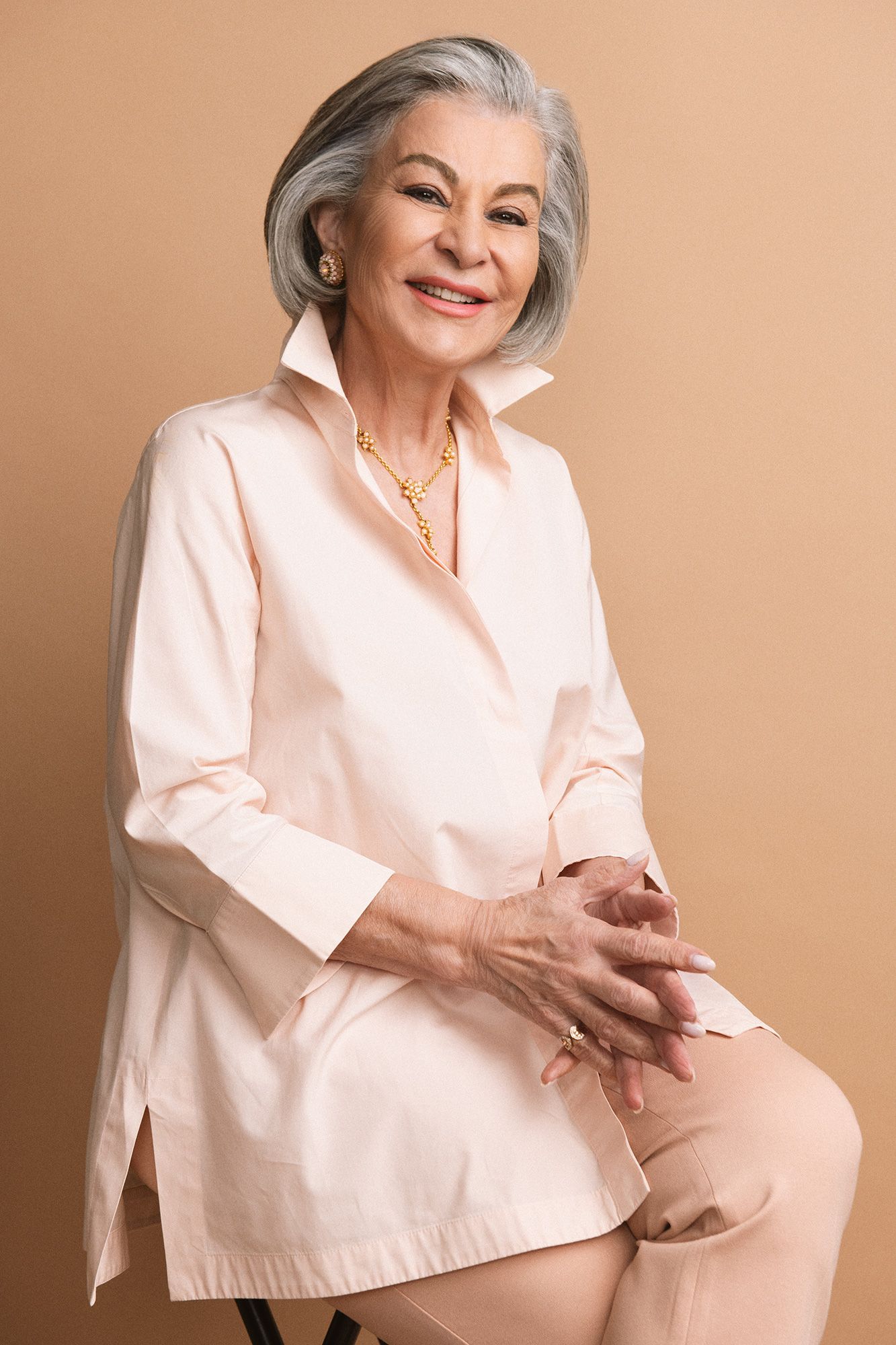 Elegant senior woman with gray hair smiling, wearing a light pink blouse and gold necklace, seated against a beige background.
