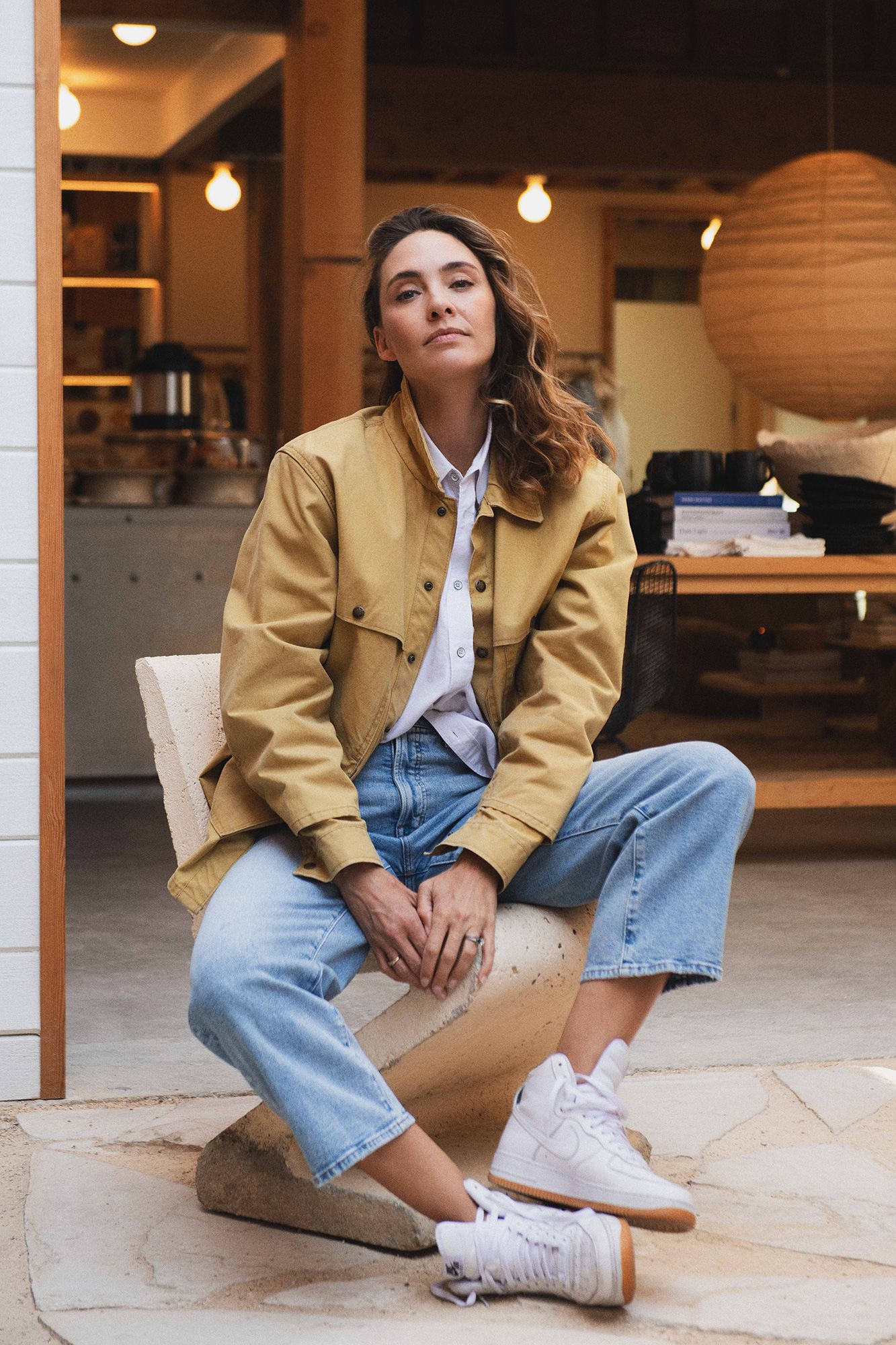 Woman in yellow jacket, jeans, and white sneakers sitting on a stone seat in a modern retail space.