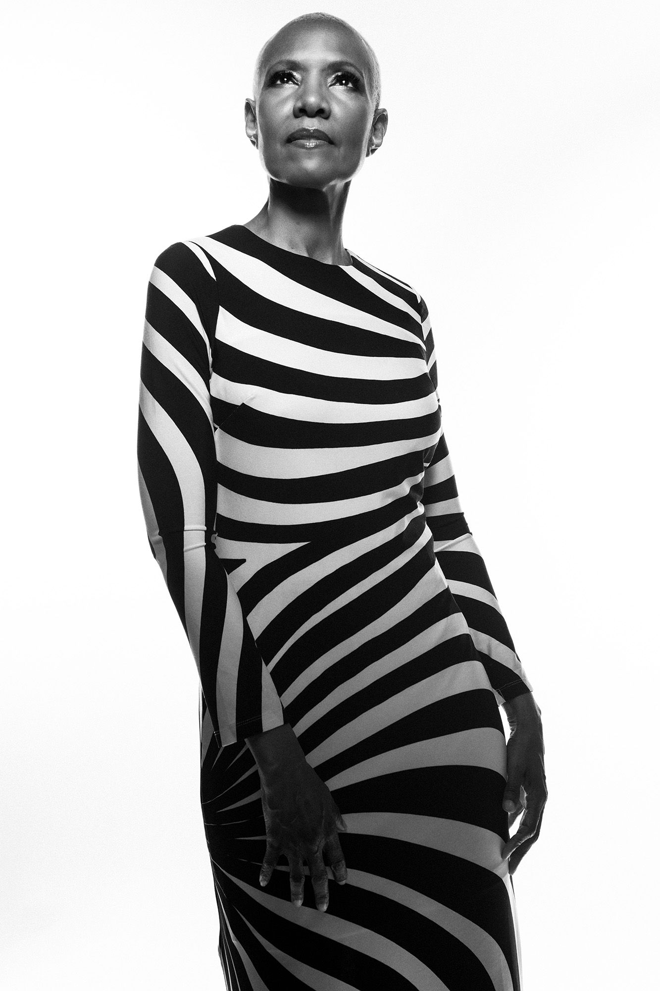 Black and white photo of a bald woman in a striped bodycon dress posing gracefully against a white background.