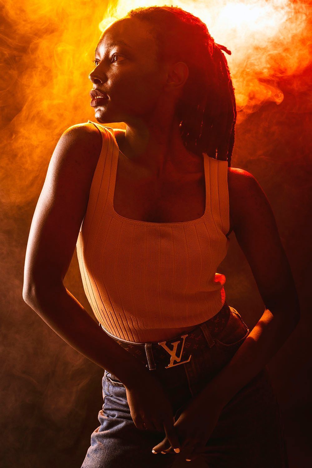 Young woman with dreadlocks in cream tank top and Louis Vuitton belt posed in orange smoky light.