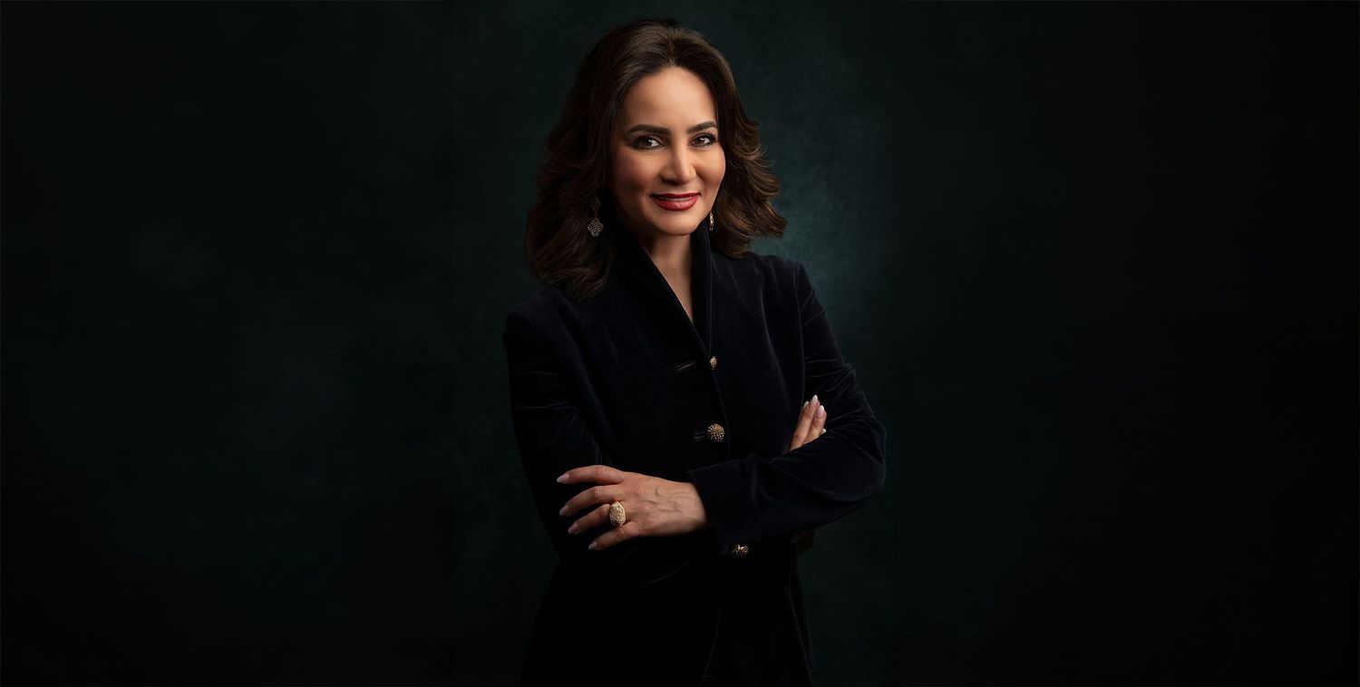 Professional woman in black blazer smiling with arms crossed against dark green studio background.