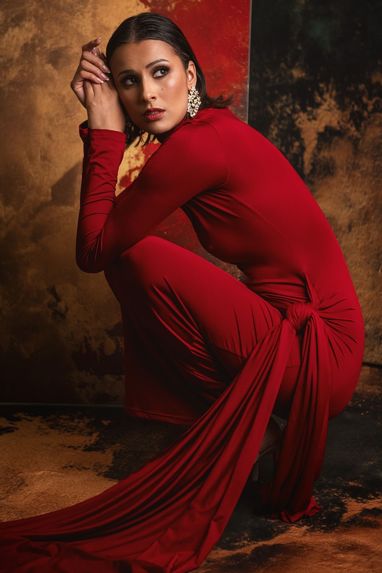Woman in red long-sleeve jumpsuit crouching against textured background.