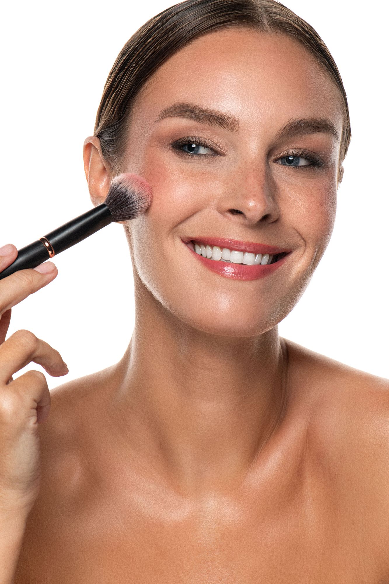 Smiling woman applying blush with makeup brush on cheekbone against white background.