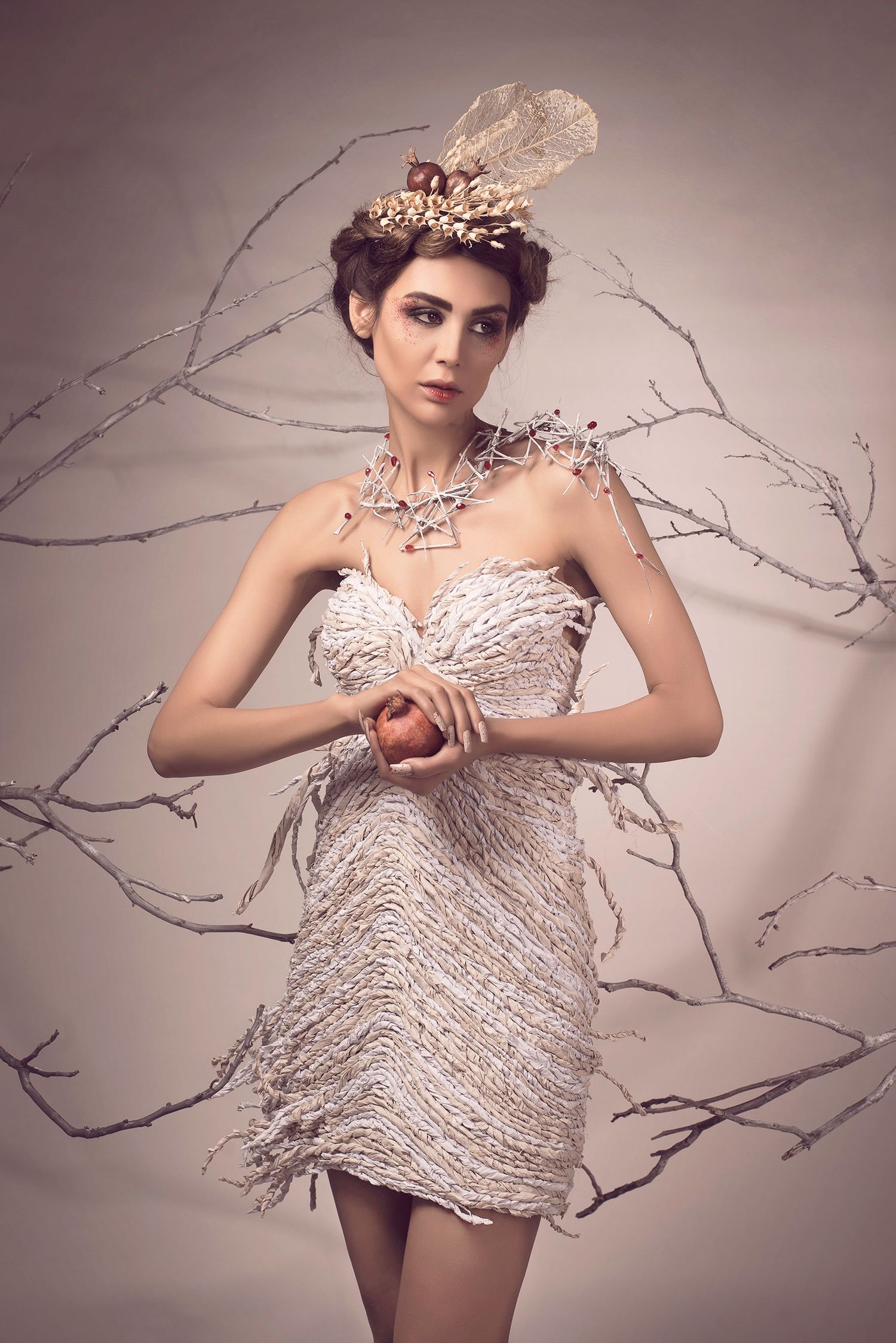 Model in white feathered mini dress holds apple, wearing ornate crown headpiece, posed among bare branches.