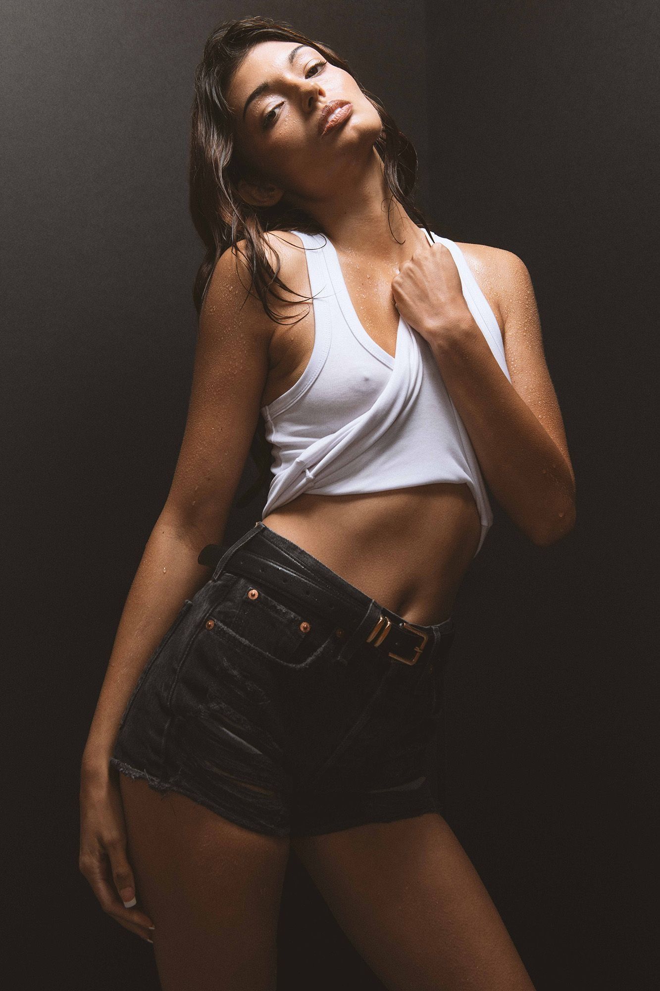 Woman in white crop top and denim shorts posing with head tilted back in dramatic studio lighting.
