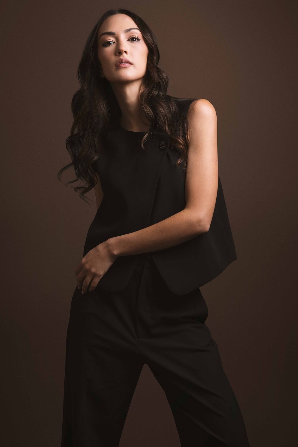 Elegant woman in black sleeveless asymmetric top and wide-leg pants on brown background.