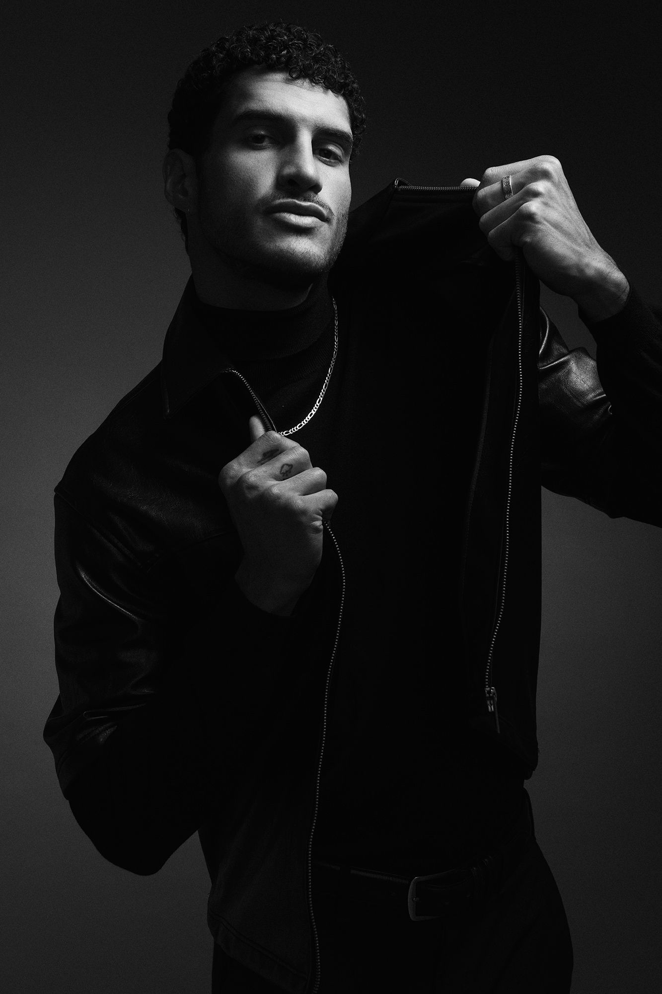 Black and white portrait of man in leather jacket holding collar with chain necklace.