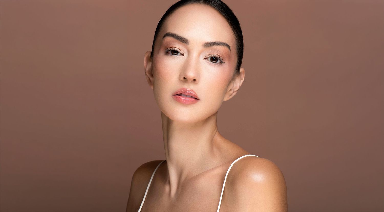 Woman with sleek updo wearing white strap top showcasing natural glowing skin against warm brown background.