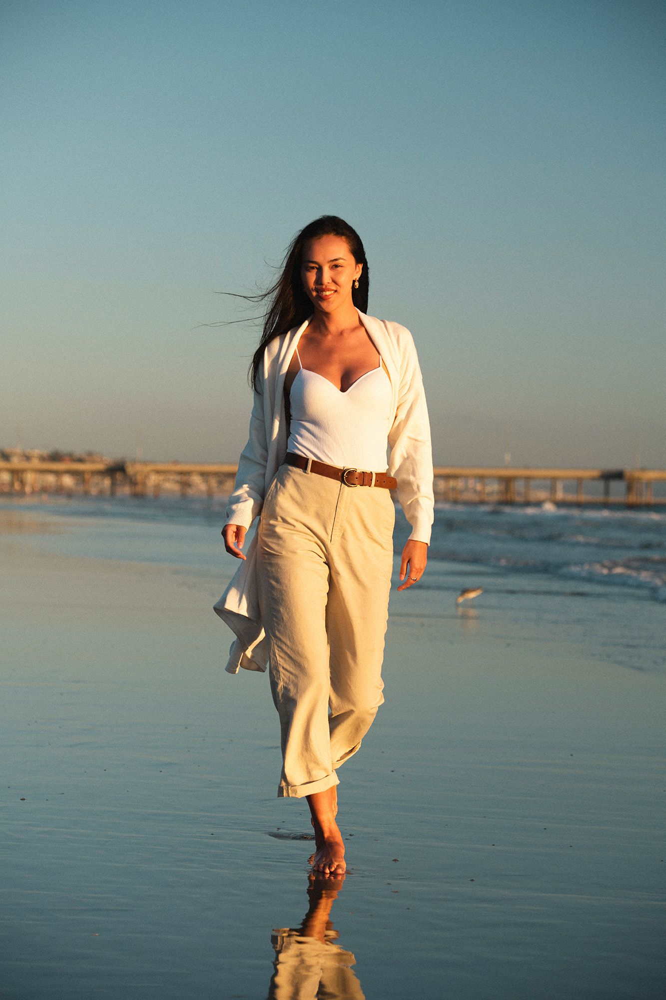 Woman in white outfit walking barefoot on a sunny beach with a pier in the background.