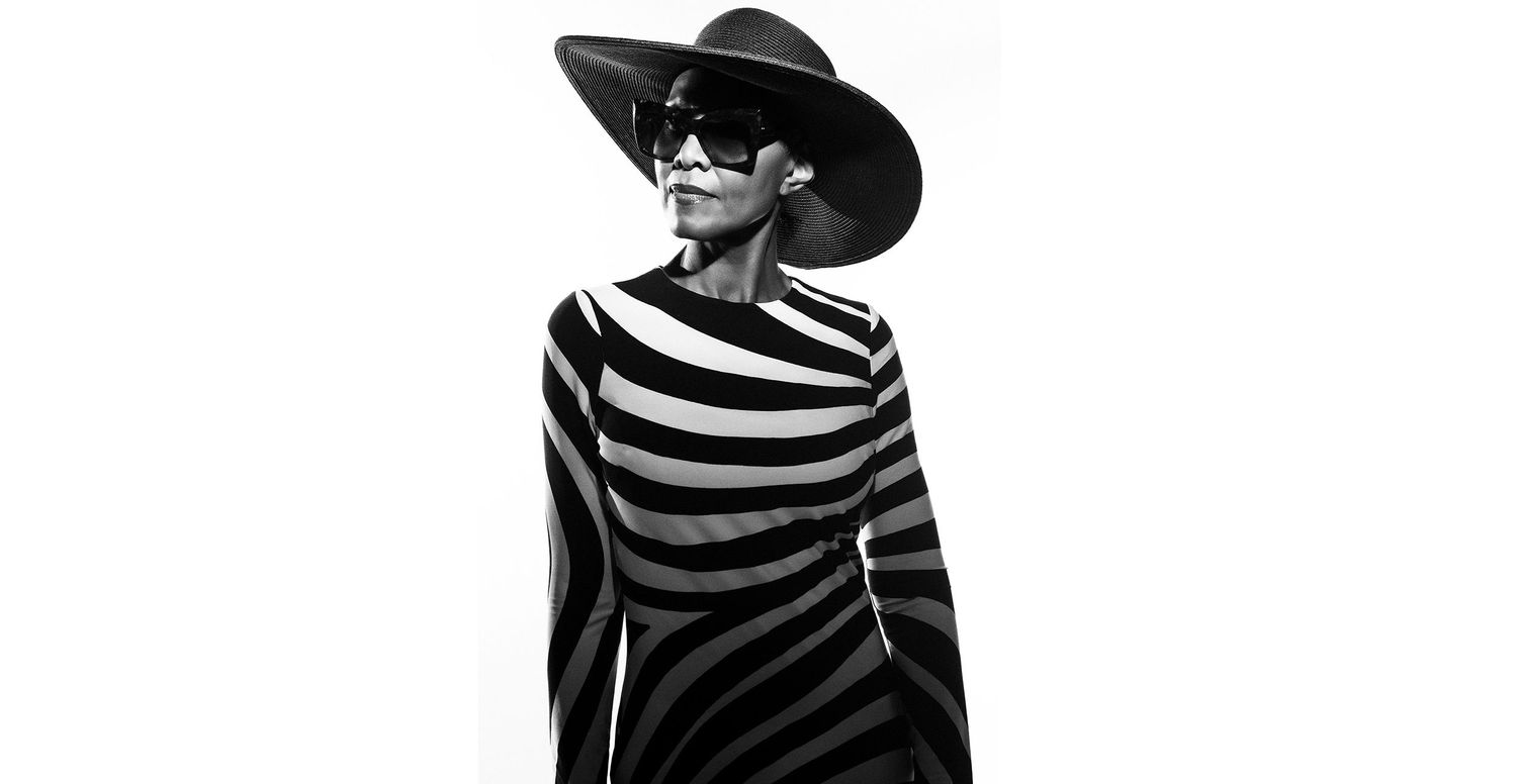 Black and white photo of a model in striped dress and wide-brim hat with sunglasses striking a bold pose.