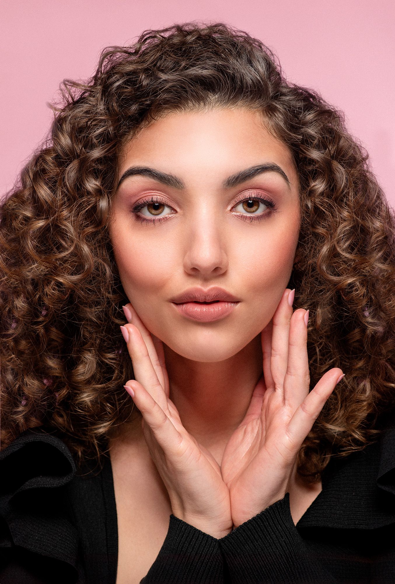 Young woman with voluminous curly brown hair and pink makeup posing against a pink background.