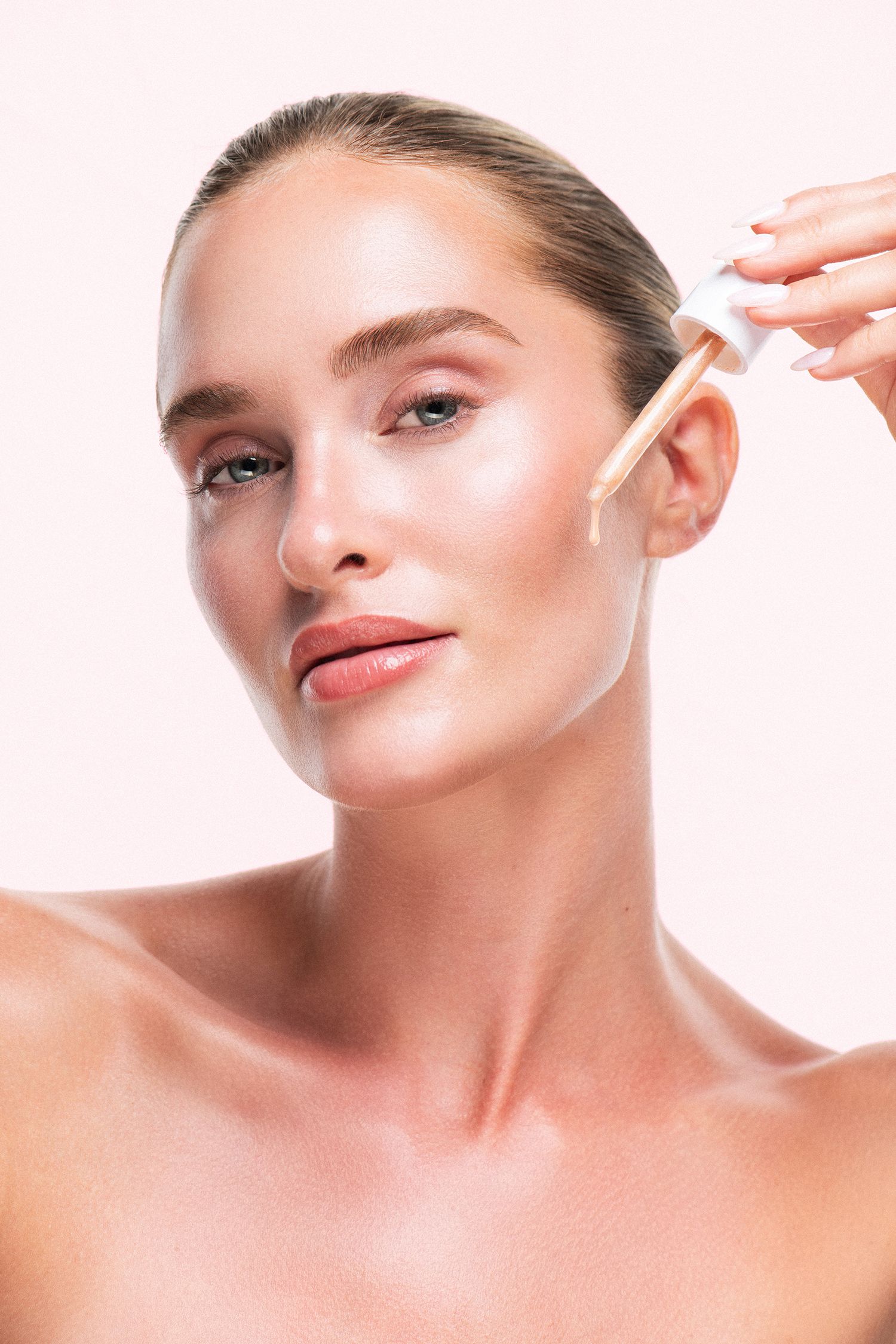 Woman applying skincare serum with dropper to glowing complexion, showcasing radiant healthy skin against soft pink background.