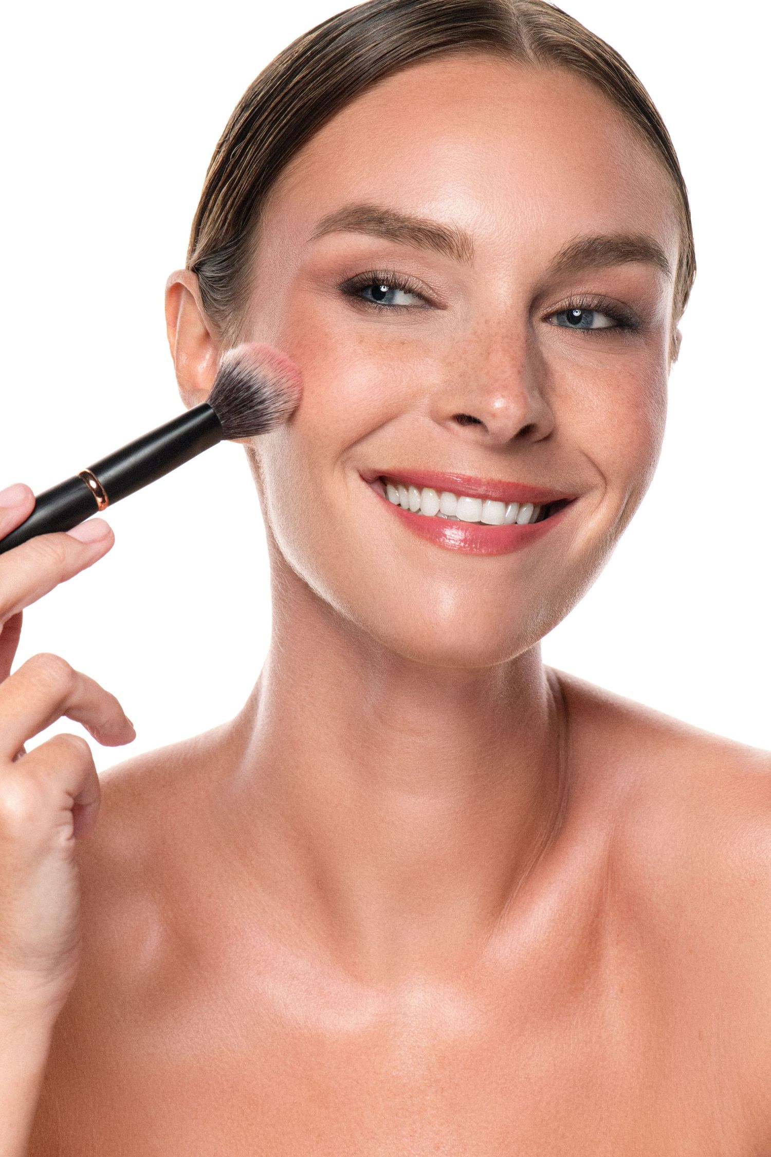 Smiling woman applying blush with makeup brush on white background, showcasing flawless skin and natural beauty look.