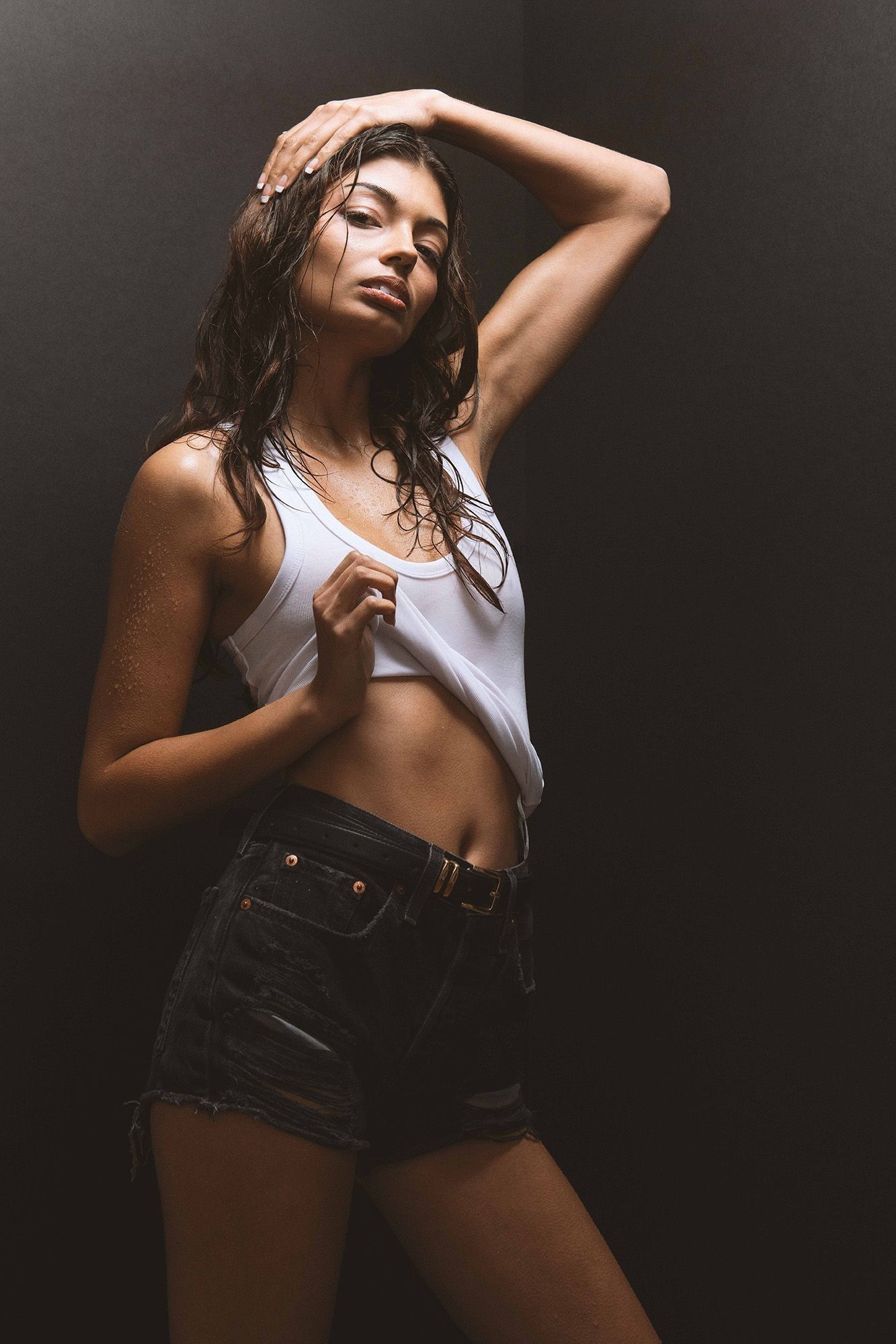 A young woman in a white crop tank top and denim cutoff shorts poses against a dark background.